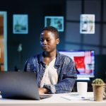 African American female student sitting at a desk typing on a digital laptop. Focused black freelancer utilizing modern technology at home office with her personal portable computer.